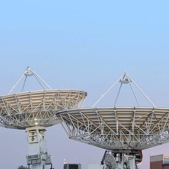 Two large, white gateway SATCOM antennas pointed at a light blue sky at dusk